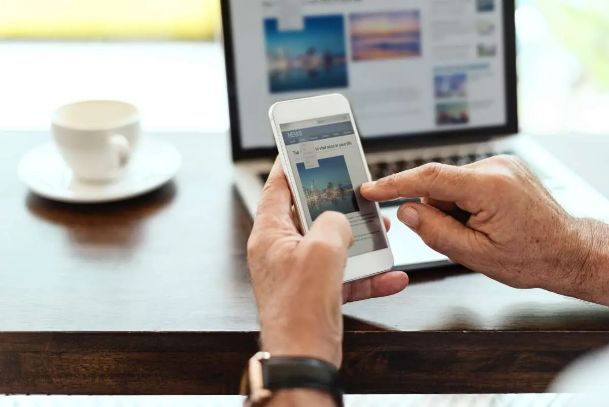 Hands holding a smartphone that shows a news app, with a laptop and a coffee cup on a wooden table