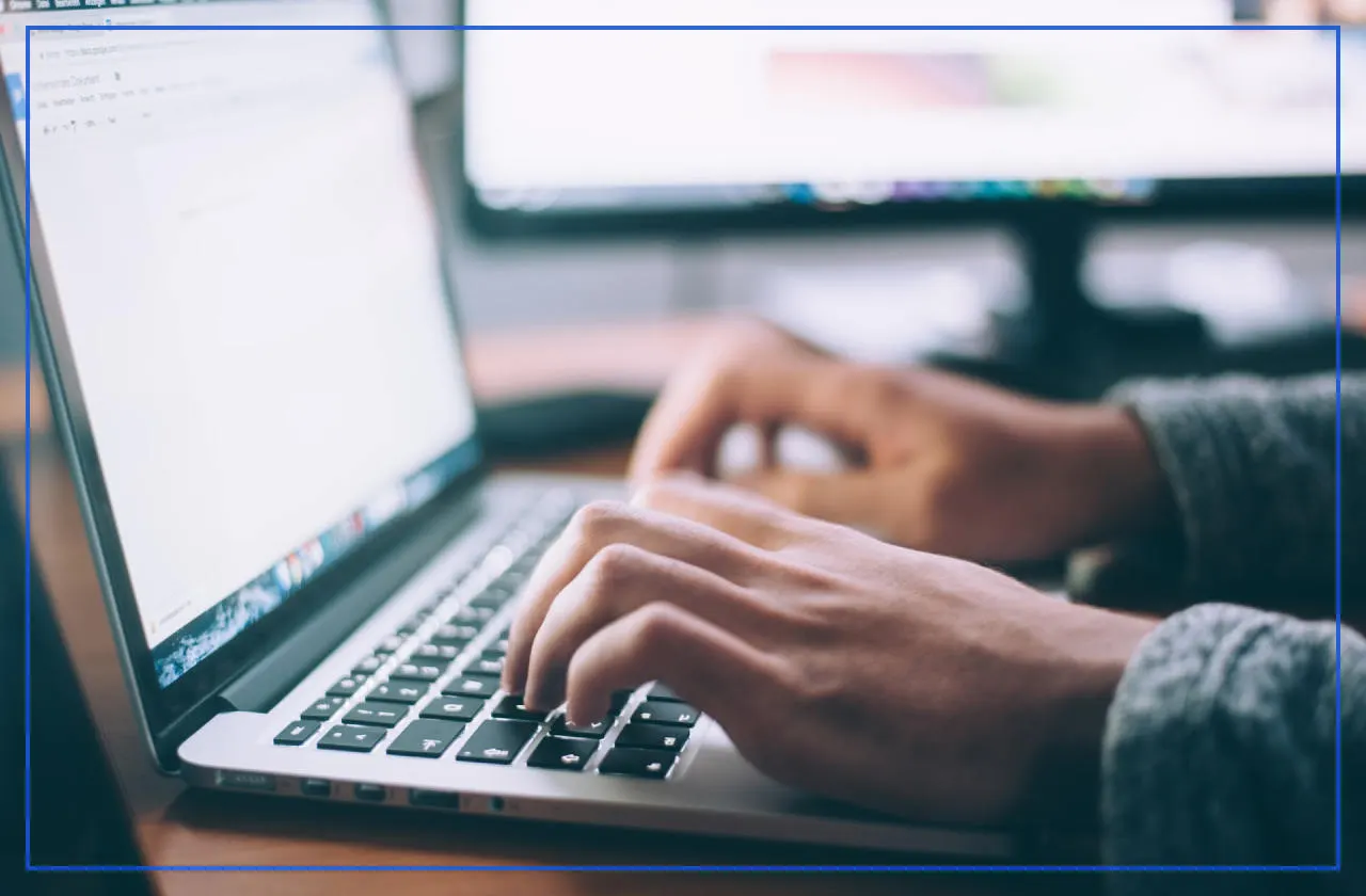 Hands typing on a laptop keyboard in front of a desktop monitor
