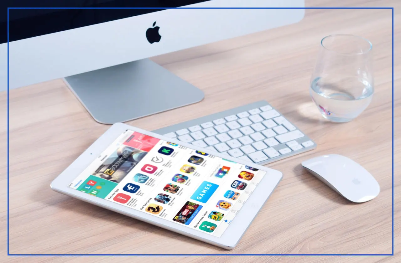 iPad showing an app store next to an iMac keyboard, a mouse and a glass of water on a wooden desk