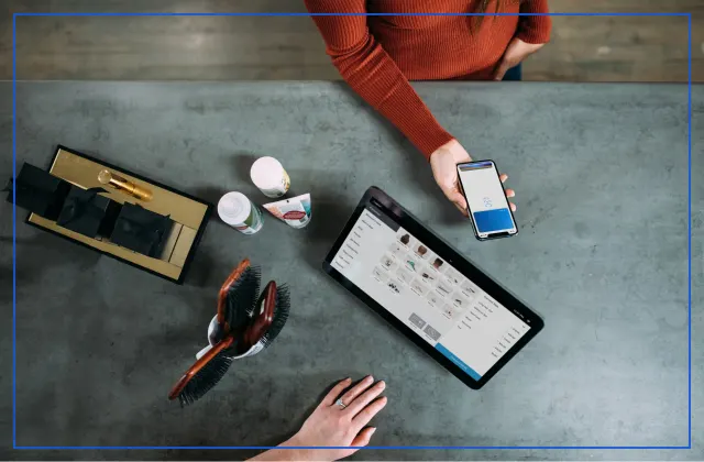 Shop counter seen from above, with a customer holding a smartphone next to a tablet showing a product catalog