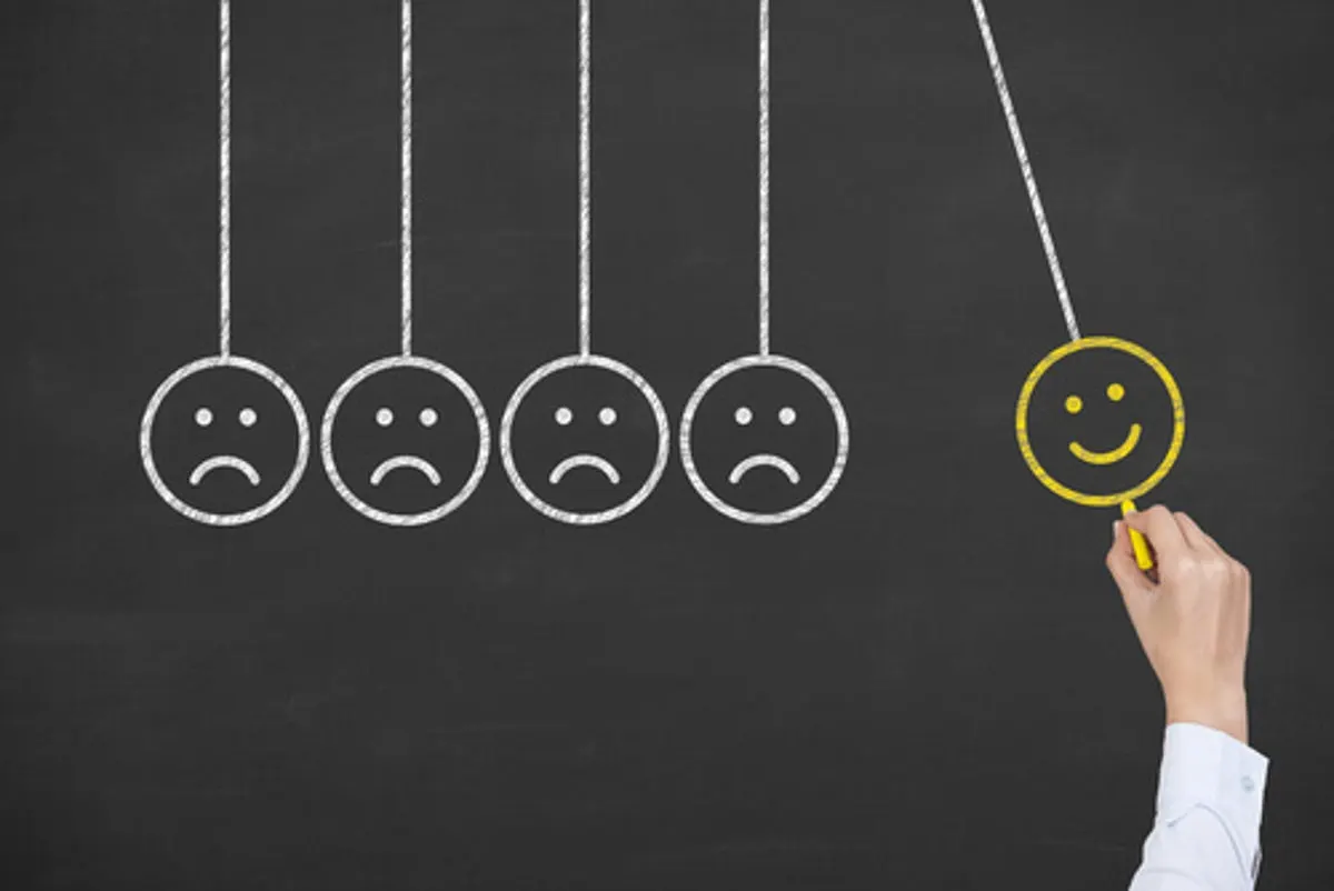 Hand drawing a yellow smiling face next to four sad faces on a chalkboard, arranged like a Newton’s cradle
