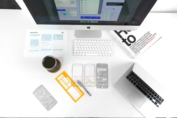 Designer’s desk with printed wireframes, mobile screen sketches, UI stencils, a desktop computer and a laptop