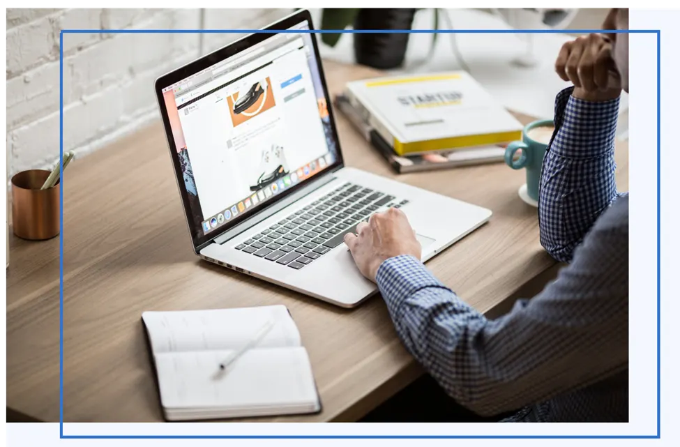 Person in a checked shirt browsing an online store on a laptop at a wooden desk