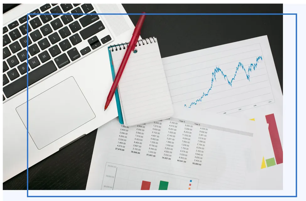 Laptop, notepad with a red pen, and printed charts and tables on a dark desk