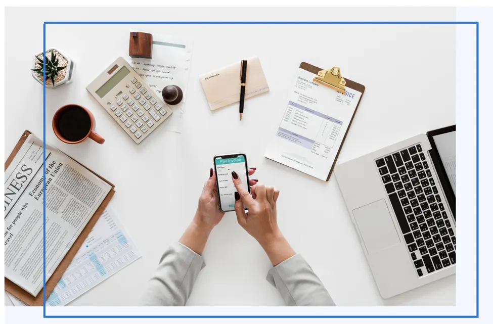 Hands holding a smartphone with a Pay Invoice screen, surrounded by a calculator, an invoice and a laptop