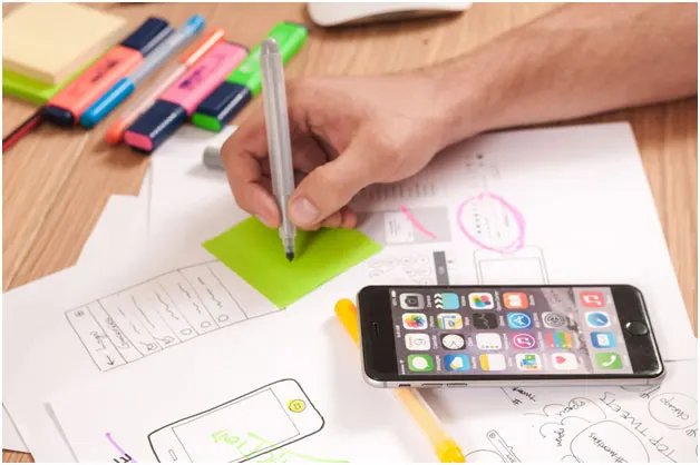 Designer sketching mobile app wireframes on paper next to a smartphone and colored markers