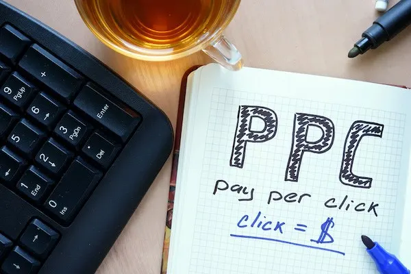 Notebook with the words PPC, pay per click and click = $ next to a keyboard and a cup of tea