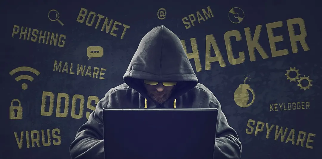 Hooded hacker at a laptop surrounded by words such as phishing, malware, DDoS, virus and spyware