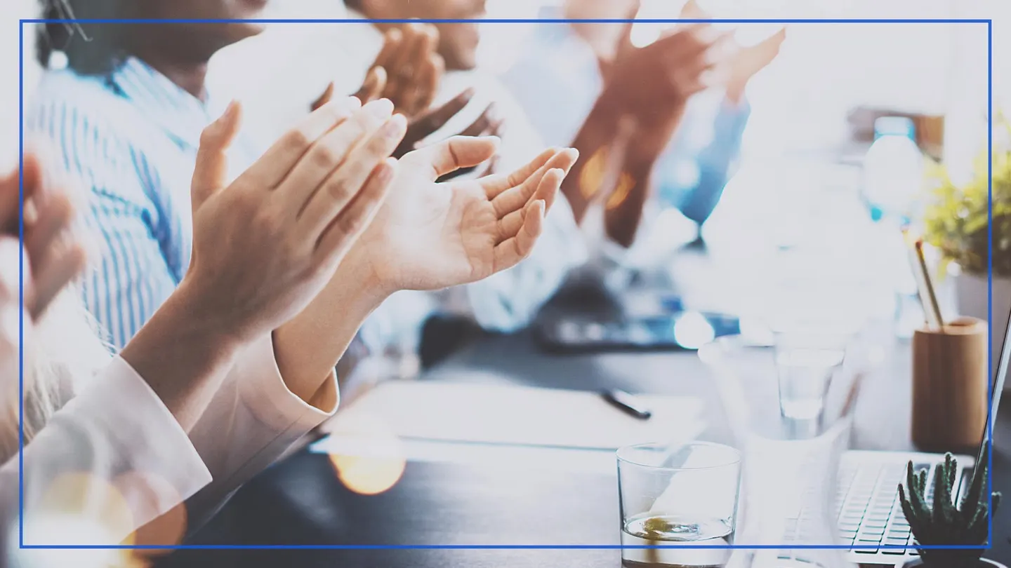 Business people applauding around a meeting table with a laptop and a glass of water