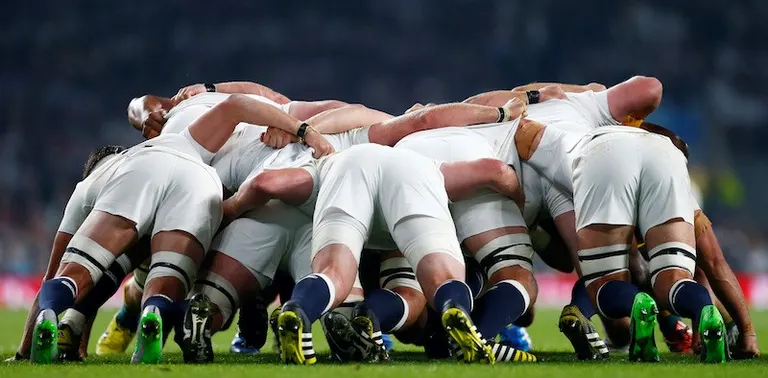 Rugby players in white jerseys locked together in a scrum on the pitch